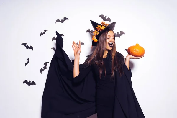 Jeune fille rieuse avec du rouge à lèvres noir sous la forme d'une sorcière pour Halloween, son manteau noir voltigeant, tenant une citrouille dans ses mains — Photo