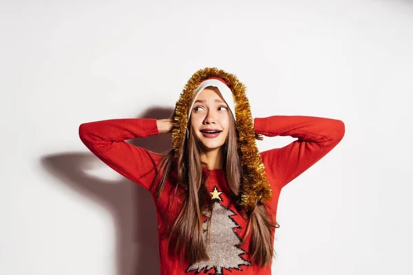 Giovane ragazza felice festeggia il nuovo anno e il Natale, in un berretto rosso, maglione festivo e fili di lame d'oro — Foto Stock