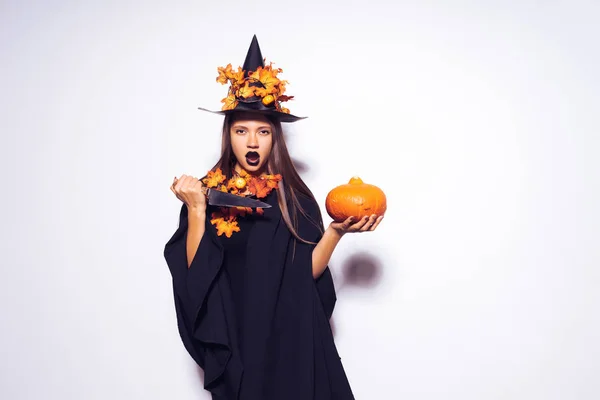 Una giovane ragazza a forma di strega ad Halloween, con un cappello nero, ornato di foglie gialle, tiene in mano un coltello e una zucca — Foto Stock