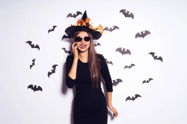 Una giovane ragazza sotto forma di strega ad Halloween, indossando un cappello nero, decorato con foglie gialle, si erge sullo sfondo di disegni di pipistrelli neri. — Foto Stock