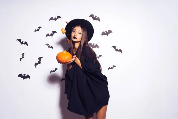 Una giovane ragazza gotica festeggia Halloween con un cappello da strega nera, tiene una zucca arancione — Foto Stock