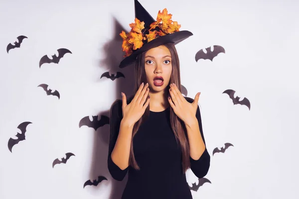 Een jonge verrast gotische meisje in een zwarte heks hoed, viert halloween. tegen de achtergrond van tekeningen van vleermuizen — Stockfoto
