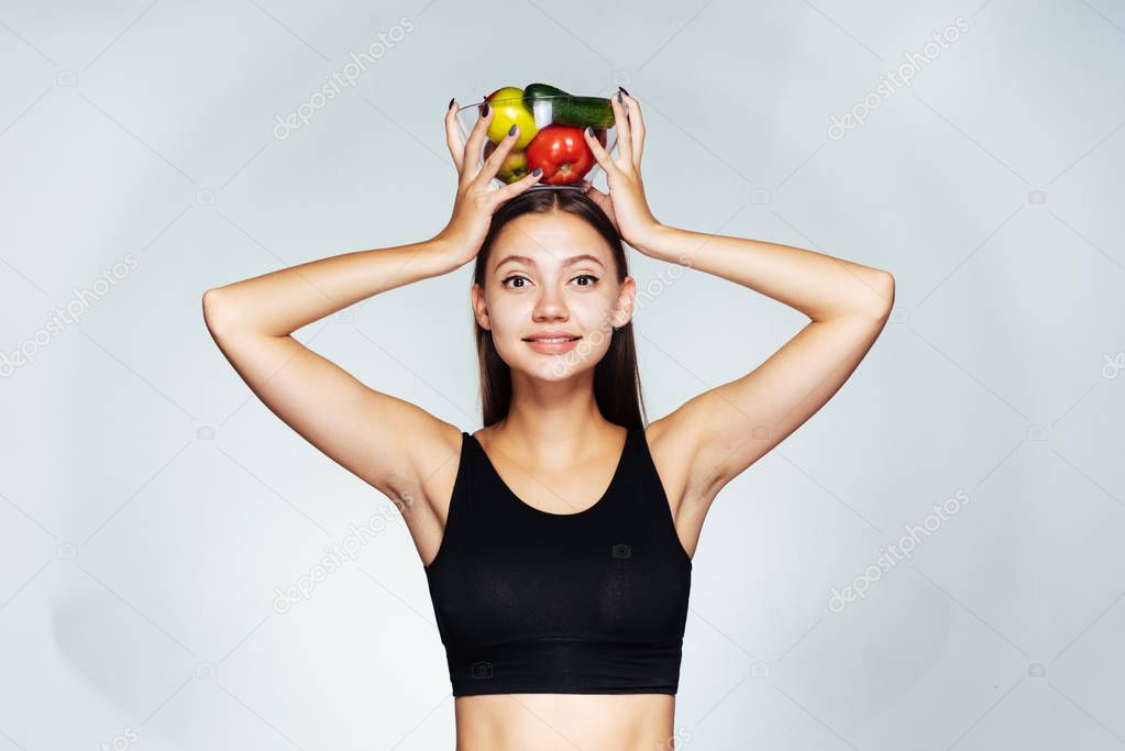 a young sundry girl watches her figure, holds a plate with useful vegetables and fruits over her head, smiles
