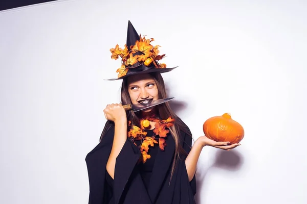 Joven chica gótica riendo en la imagen de una bruja en Halloween, sosteniendo un cuchillo y una calabaza en su mano —  Fotos de Stock