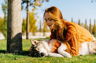 güzel kızıl saçlı kız yeşil çimenlerin üzerinde yatıyor ve köpeğini parkta oynuyor. Mükemmel güneşli bir sonbahar günü.