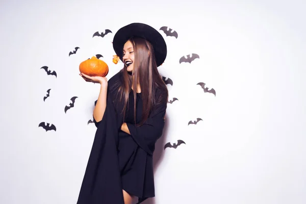 Uma menina gótica bonita nova na forma de uma bruxa em halloween, um chapéu preto grande decorado com folhas amarelas, segurando uma abóbora — Fotografia de Stock