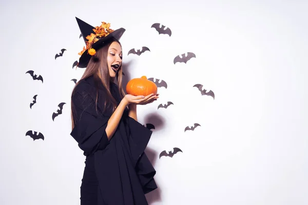 Een prachtige gotische meisje in de vorm van een heks in halloween, een grote zwarte hoed versierd met gele bladeren, houden een pompoen — Stockfoto