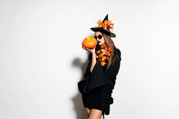 Une jeune belle gothique en forme de sorcière à Halloween, un grand chapeau noir orné de feuilles jaunes, tenant une citrouille — Photo