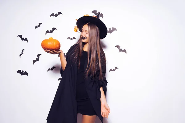 Une jeune belle gothique en forme de sorcière à Halloween, un grand chapeau noir orné de feuilles jaunes, tenant une citrouille — Photo