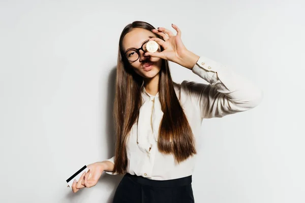 Una buena chica con gafas está sosteniendo una tarjeta bancaria en su mano y adjuntando una moneda de oro a su ojo. Bitcoins, criptomoneda, dinero electrónico . — Foto de Stock