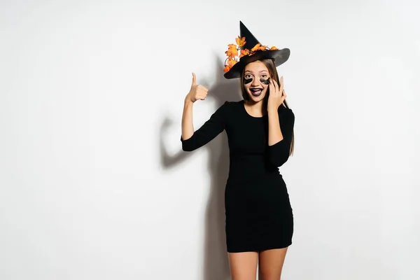 Bruja de Halloween en un terrible traje y sombrero muestra los pulgares hacia arriba y se ríe. La chica iba a la fiesta de Halloween. . —  Fotos de Stock