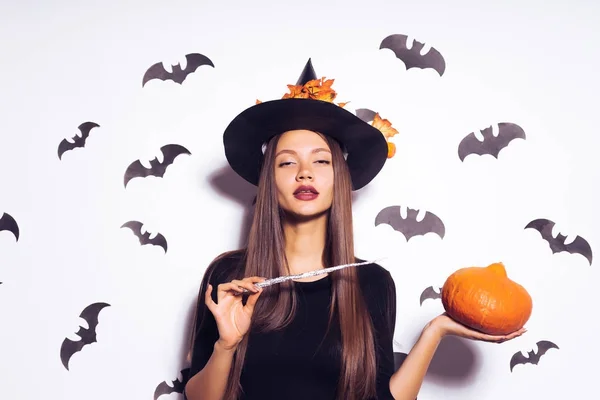 Uma bruxa íngreme evoca uma abóbora com sua varinha mágica e olha para a câmera. Bruxa em um vestido preto e chapéu com folhas caídas. Menina na festa de Halloween. Roupa de Halloween, férias . — Fotografia de Stock