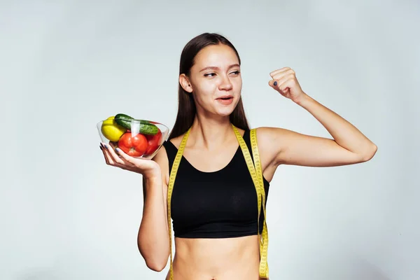 En sportig tjej håller en skål med grönsaker och visar hur stark hon är. Vacker kropp, rätt kost, hälsosam mat, fitness. — Stockfoto