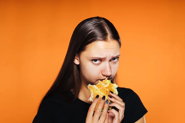 Üzgün kız kameraya bakar ve turuncu bir arka plan üzerinde izole bir hamburger yiyor — Stok fotoğraf