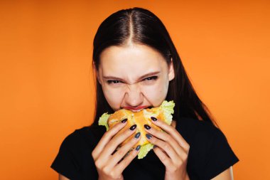 Kız iştahla hamburger yiyor ve öfkeyle kameraya benziyor. Turuncu zemin üzerine izole lezzetli yemekler