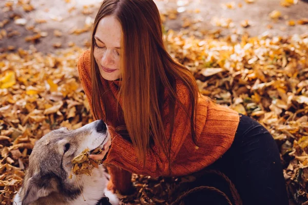 Young red-haired girl is playing with her dog in the park, around the fallen yellow leaves — Stock Photo, Image