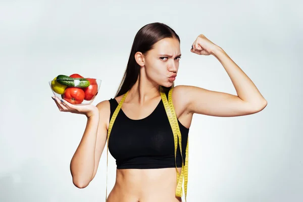 Una joven atlética observa su figura, sostiene un plato con verduras en las manos y demuestra su bíceps —  Fotos de Stock