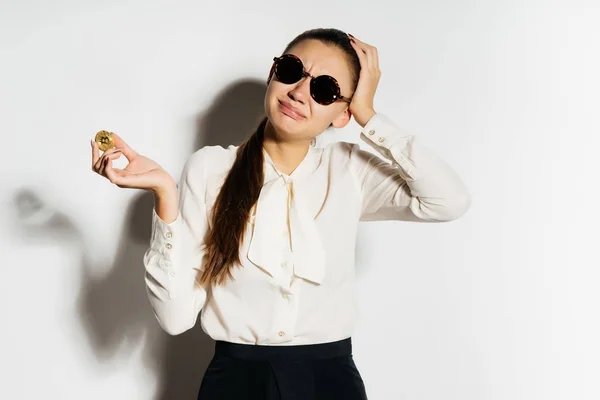 Chica extraña con gafas y una blusa blanca sosteniendo una moneda de oro y mirando a la cámara, criptomoneda, bitcoins, aislado sobre un fondo blanco — Foto de Stock