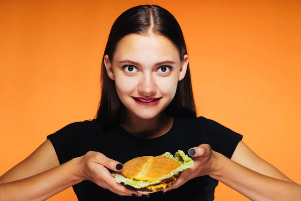 Ta dívka drží chutný hamburger a dychtivě při pohledu na fotoaparát. Izolované na oranžové pozadí. — Stock fotografie