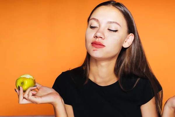 Hermosa chica come una manzana y linda mira hacia abajo. Comida saludable, comida sabrosa, aislada sobre fondo naranja . — Foto de Stock