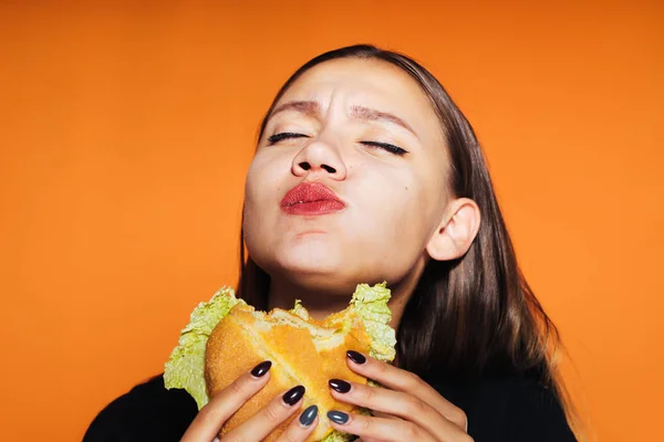 La chica come codiciosamente una hamburguesa. sabrosa comida, aislada sobre fondo naranja —  Fotos de Stock