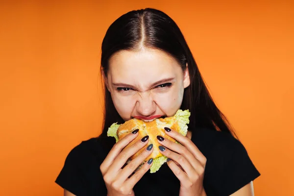 Dívka hltavě jí hamburger a zlostně se dívá na kameru. chutné jídlo, izolované na oranžové pozadí — Stock fotografie