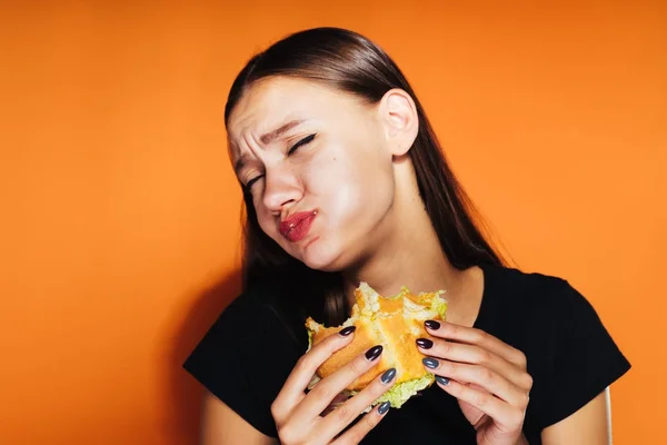La fille affamée mange son hamburger. Nourriture très savoureuse, isolée sur un fond orange — Photo