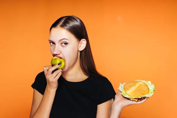 Mladá dívka chce zhubnout, jí zdravé jablko a v ruce drží škodlivé burger — Stock fotografie