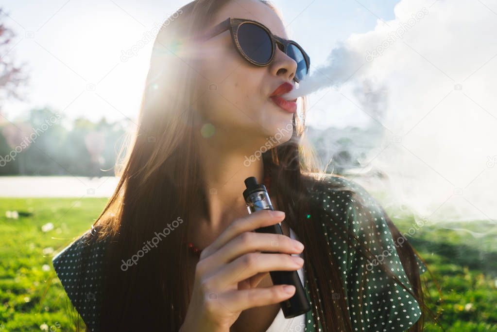 Happy fashion smiling Woman in sunglasses smoking vape on street, smoke
