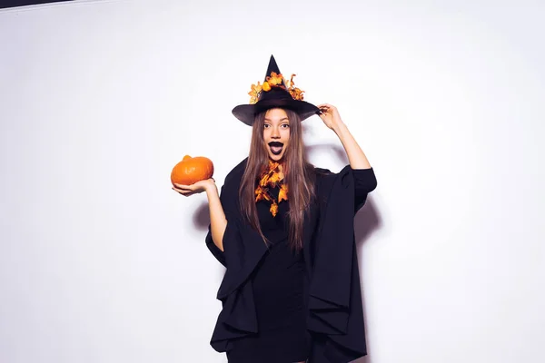 Mladé sexy tajemná dívka oblečená v čarodějnice, slaví halloween, velkým černým kloboukem, zdobí žluté listy a drží dýně — Stock fotografie