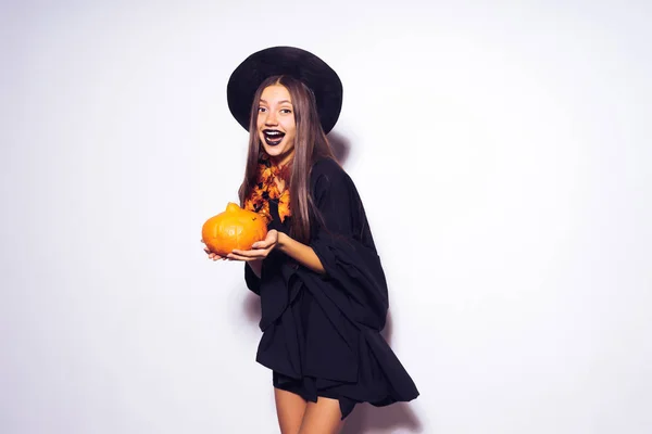 Joven chica gótica sexy en la forma de una bruja en Halloween, en un gran sombrero negro, sosteniendo una calabaza en sus manos — Foto de Stock