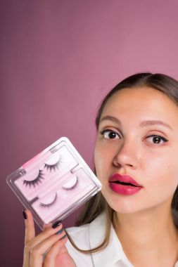 Büyük gözlü güzel bir kız yanlış eyelashes onun elinde tutar. Yüz yakın çekim, pembe bir arka plan üzerinde izole