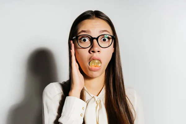 Молодая современная успешная деловая женщина в очках держит золотой биткойн во рту и выглядит удивленной — стоковое фото