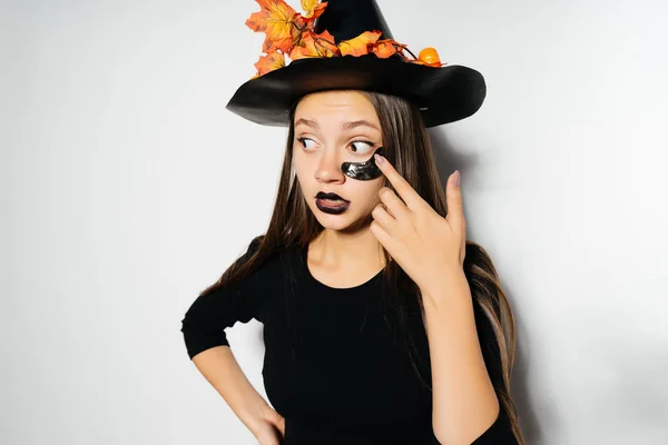Una joven mujer gótica sexy en forma de bruja en Halloween, con un gran sombrero negro, adornado con hojas amarillas, parece sorprendida —  Fotos de Stock