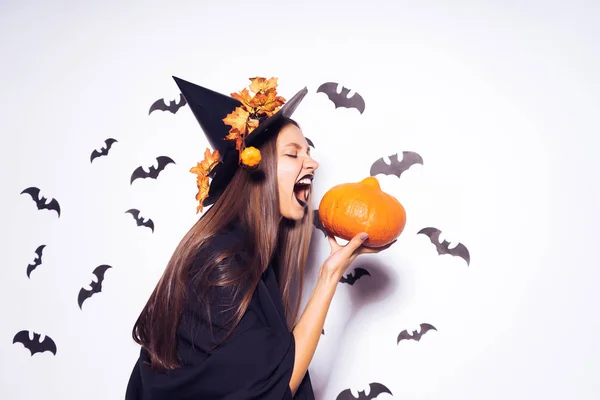a young sexy gothic woman in the form of a witch in halloween, in a big black hat, adorned with yellow leaves, holds a pumpkin and screams at her
