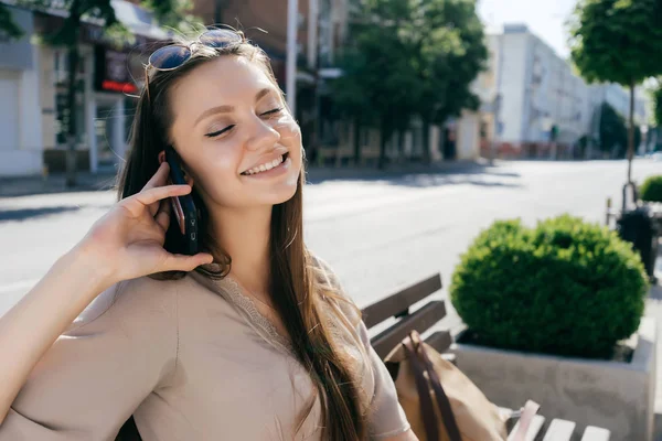 Chica encantadora en un paseo se sienta en un banco y sonriendo hablando por teléfono — Foto de Stock