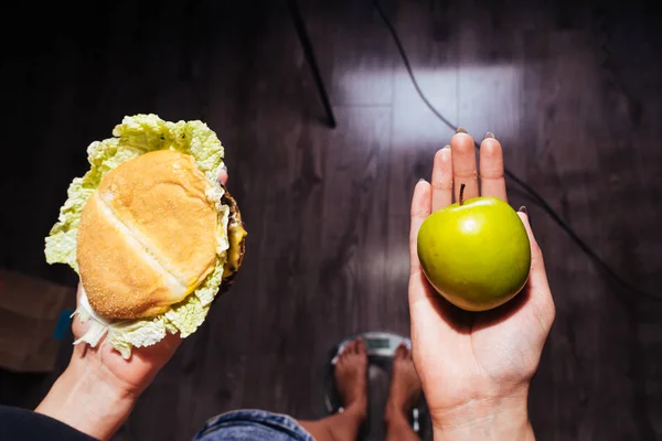 Ein junges schönes Mädchen beobachtet ihre Gesundheit und Figur, wie sie auf der Waage steht und einen nützlichen Apfel und einen schädlichen Burger in der Hand hält — Stockfoto