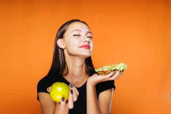 Uma menina goza de um hambúrguer em suas mãos segurando em outra maçã — Fotografia de Stock