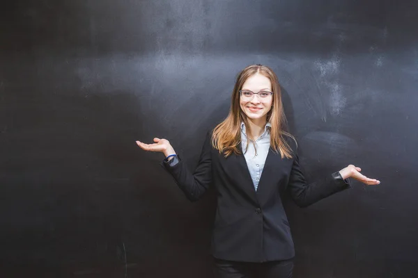 Ein nettes Mädchen mit Brille und dunklem Büroanzug steht an der Tafel, lächelt und breitet die Arme zur Seite aus — Stockfoto