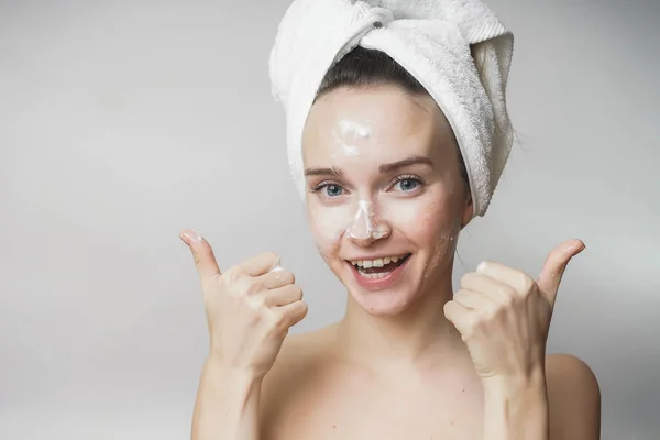 Femeie amuzantă într-un prosop pe cap fericit curăță pielea cu spumă, arată degetele mari — Fotografie, imagine de stoc