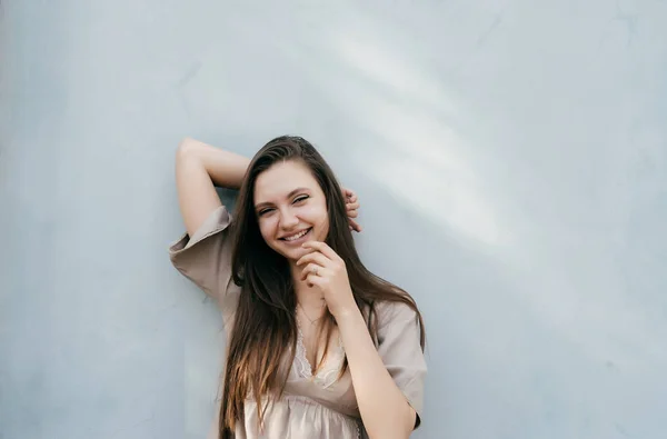 Linda chica de pelo largo en vestido beige de pie junto a la pared lindo sonriendo y mirando a la cámara — Foto de Stock
