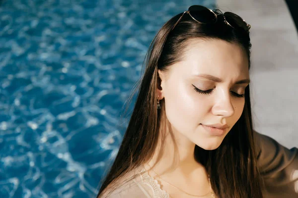 Hermosa y encantadora chica se sienta junto a la piscina con los ojos cerrados y disfruta de un cálido día de verano — Foto de Stock
