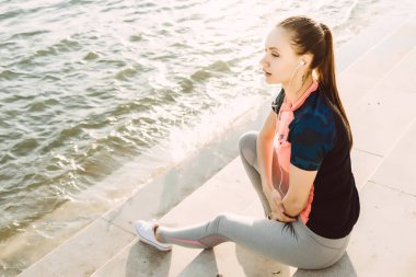 Suyun kenarında oturan kulaklıklar, müzik dinlemek, Irmak, akşamları koşan bir genç atletik kız