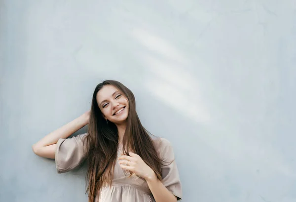 Joven hermosa chica en vestido beige y pelo largo posando contra la pared y sonriendo — Foto de Stock