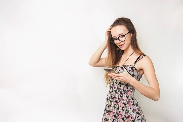 Молодая милая девушка в очках и платье улыбаясь и глядя в ее смартфон — стоковое фото