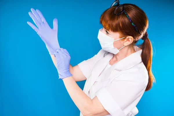 Adult rödhårig kvinna läkare i vit labbrock sätter på blå medicinska handskar — Stockfoto