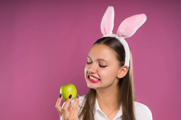 En rolig flicka med en fälg i form av hares öron håller i sin hand ett grönt äpple — Stockfoto