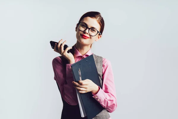Junges modernes süßes Mädchen mit Brille in grauer Büroweste lächelt, hält einen Ordner mit Dokumenten und Telefon in der Hand — Stockfoto