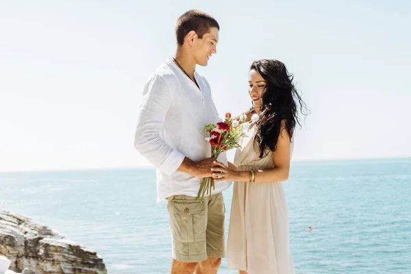 Два любовника стоят на скале, парень дарит девушке цветы, романтика в отпуске — стоковое фото