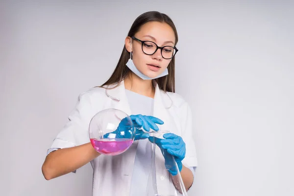 En ung flicka med glasögon och en medicinsk morgonrock, iklädd blå medicinska handskar gör experiment med kemikalier i kolvar — Stockfoto